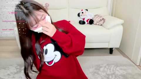 Media: Video of a woman in a red Mickey Mouse sweater, wearing a white face mask, kneeling on a white rug, in a modern living room with a white couch and stuffed toys.