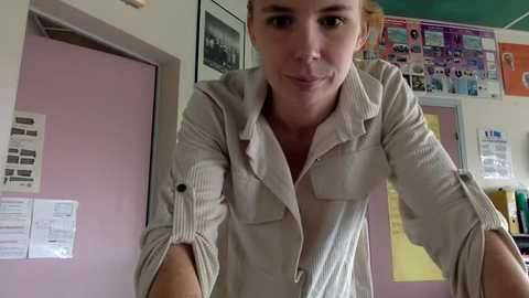 Media: Video of a light-skinned woman with short blonde hair, wearing a beige button-down shirt, standing in a classroom with pastel walls and bulletin boards, smiling slightly.