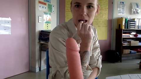 Media: Video of a blonde woman in beige shirt, holding a large pink dildo, in a classroom with yellow bulletin boards and shelves.