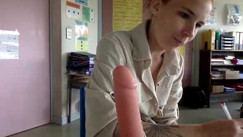 Media: Video of a light-skinned woman with brown hair, wearing a beige blouse, in a classroom. She holds a large, realistic, erect, pink dildo in front of her, with a small, red pen in her other hand. Background includes colorful posters, books, and shelves.