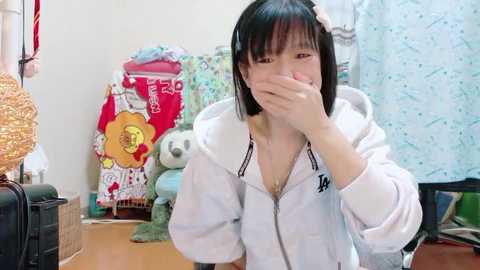 Media: A young East Asian woman with straight black hair and a white hoodie, covering her face with one hand, sits on a wooden floor in a cluttered room with a teddy bear, a colorful blanket, and a patterned curtain in the background.