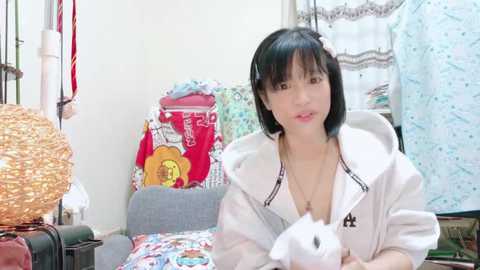 Media: A video of an Asian woman with short black hair, wearing a white hoodie, holding a white cat, in a cluttered, colorful room with a wicker basket, colorful blankets, and a window with white curtains.