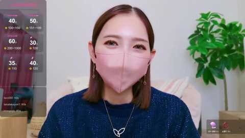 Media: Video of an East Asian woman with straight, shoulder-length brown hair, wearing a pink face mask, dark blue sweater, and a butterfly necklace, set against a plain background with a potted plant and a weather forecast overlay.