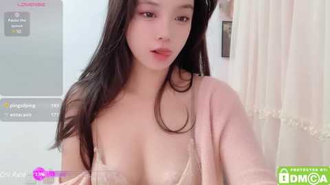 Media: Video of an Asian woman with long black hair, wearing a pink cardigan and a lace bra, looking at the camera with a neutral expression. Background features a white curtain and a gray wall with social media icons.