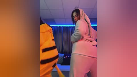 Media: Video of a young woman in a white onesie with a tiger costume, standing in a dimly lit room with blue LED strip lights, and a bed in the background.