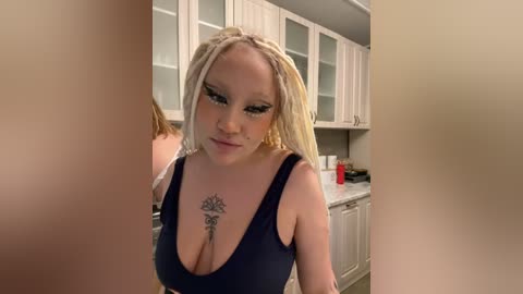 Media: Video of a blonde woman with large breasts wearing a low-cut black top, standing in a modern kitchen with white cabinets and a red container.