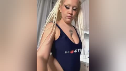 Media: Video of a plus-sized woman with long blonde dreadlocks, wearing a dark blue swimsuit, in a kitchen. She has a tattoo on her chest and is looking downward.