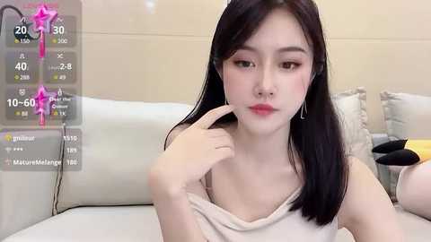 Media: Video of an East Asian woman with long black hair, fair skin, and a slender physique, wearing a beige tank top, sitting on a white couch, with a virtual overlay showing her age, height, and weight.