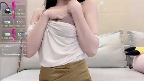 Media: A video of a slim, light-skinned woman with long black hair, partially covered by a white cloth, wearing a beige skirt. Background includes a white bed with pillows and a stuffed toy.