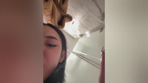 Media: Video of a young woman with long black hair and fair skin, partially visible from the neck up, wearing black eyeliner and mascara, standing in a bathroom with beige walls, a tiled ceiling, and a towel hanging above her head.