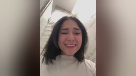 Media: A video of a young woman with straight black hair, fair skin, and a slight smile, wearing a white turtleneck, standing in a dimly lit hallway with worn gray walls.