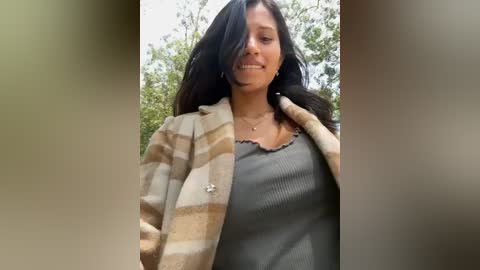 Media: Video of a woman with long black hair, wearing a beige and brown striped cardigan over a gray ribbed tank top, standing outdoors with blurred trees in the background.