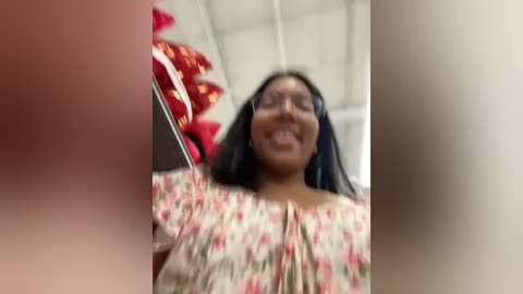 Media: Video of a smiling South Asian woman with medium skin tone, wearing a floral blouse, inside a brightly lit room with red and white decor.