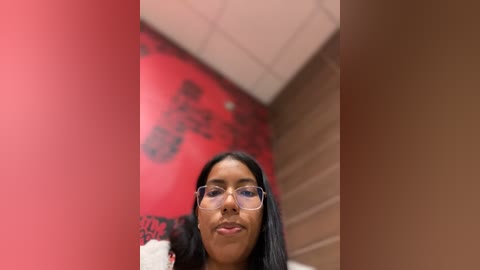 Media: A video of a young woman with long black hair, wearing glasses, standing in a red-walled hallway with a blurred mural and beige ceiling.