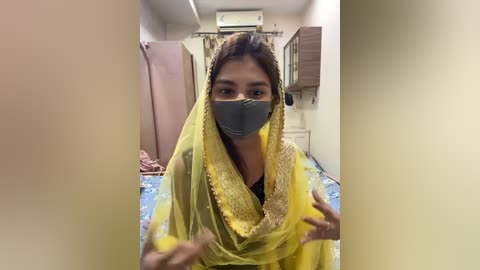 Media: Video of a woman in a yellow sari, grey face mask, and black top, standing indoors with a blue-patterned rug, white walls, and a pink curtain in the background.