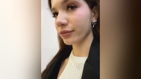 Media: Video of a young woman with light brown skin, long dark hair, and brown eyes, wearing a white knit top and a black blazer. She has small hoop earrings and a subtle makeup look. Background is plain and light-colored.