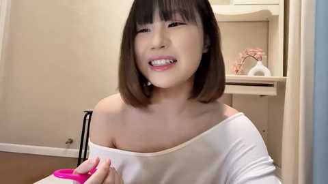 Media: Video of an East Asian woman with straight, shoulder-length brown hair, wearing a white off-shoulder top, smiling while holding a pink pen. Background features a beige wall, white kitchen cabinets, and a pink decorative item.