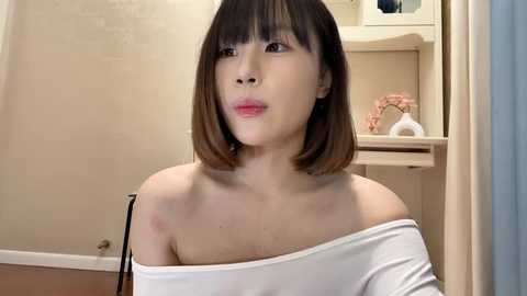 Media: A video of an Asian woman with shoulder-length brown hair and fair skin, wearing a white off-shoulder top, seated indoors. Background features a beige wall and a wooden shelf with decorative items.