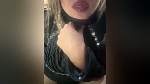 Media: Video of a woman with light skin and blonde hair, wearing a black dress with silver beads and a choker, slightly parted lips, and a hand touching her neck. Background is blurred.