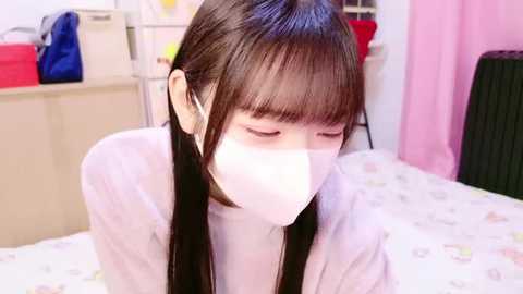 Media: A video of an Asian woman with long black hair wearing a white face mask, sitting on a bed with a white sheet, in a room with pink curtains and storage units in the background.