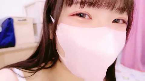 Media: A close-up video of an East Asian woman with fair skin, wearing a white face mask, brown hair, and pink eyeshadow. Background shows a white cabinet and a blue garment.