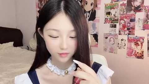 Media: Video of a young East Asian woman with long black hair, fair skin, and delicate features, wearing a frilly white choker, blue dress, and white gloves, in a cozy bedroom adorned with anime posters and a neatly made bed.