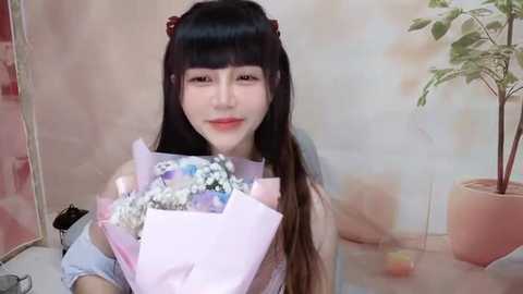 Media: Video of a smiling Asian woman with long black hair in pigtails, holding a pink bouquet with white and blue flowers. Background features a pastel-colored wall and a green plant in a pot.