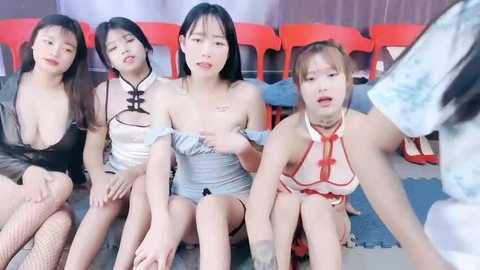 Media: Video of five Asian women in revealing, tight-fitting, semi-nude outfits, sitting on a gym mat floor, with red plastic chairs in the background.