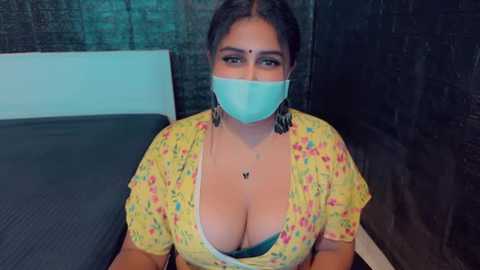 Media: Video of a South Asian woman with light brown skin, wearing a yellow floral blouse revealing ample cleavage, a teal face mask, and dangling earrings. She sits on a bed with dark sheets against a dark brick wall background.