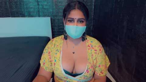 Media: Video of a South Asian woman with medium skin tone, wearing a yellow floral blouse with a low neckline revealing cleavage, a blue face mask, and a blue bra. She sits on a bed with dark bedding in a dimly lit room with a brick wall.