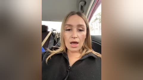 Media: Video of a blonde woman in a black jacket, with a neutral expression, sitting in a car with a blurry background.