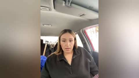 Media: Video of a woman with light brown skin, long brown hair, and wearing a black jacket, sitting in a car with a blue jacket hanging on the backseat.