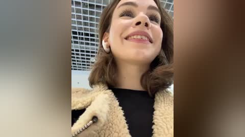 Media: A video of a young woman with fair skin, shoulder-length brown hair, and a friendly smile, wearing a black top and a beige, fuzzy jacket. The background features a grid-patterned ceiling and a blurred, brown-toned edge.