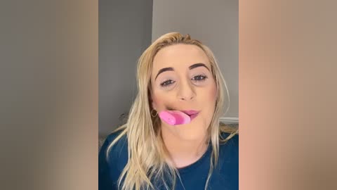 Media: Video of a blonde woman with straight hair, light skin, and dark eyebrows, wearing a dark blue shirt, licking a pink lollipop, in a plain, beige-walled room.