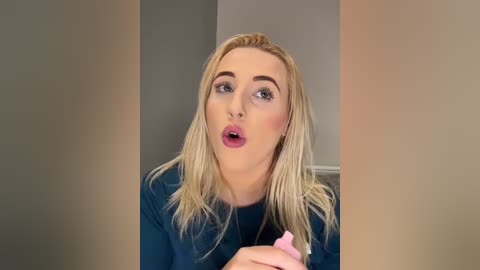 Media: Video of a fair-skinned, blonde woman with straight hair, wearing a blue shirt, and a shocked expression, holding a pink object, against a grey wall.