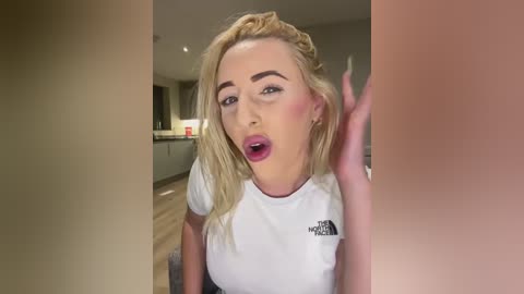 Media: Video of a blonde woman with fair skin and medium-sized breasts wearing a white t-shirt, looking surprised and raising her hand in a modern kitchen.