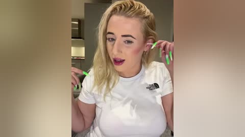 Media: Video of a blonde woman with medium skin tone, wearing a white t-shirt, playing with neon green hair extensions in a modern kitchen.