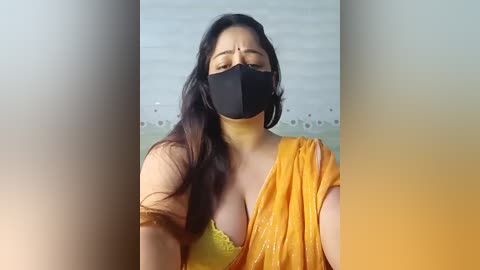 Media: Video of a curvaceous woman with medium skin tone, wearing a black mask, a yellow sari, and a yellow bra, standing indoors with blurred background.