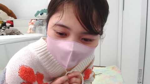 Media: A video of an Asian woman with short brown hair, wearing a pink surgical mask and a white sweater with orange patterns, seated in a bright, cluttered room with toys and a white cabinet in the background.