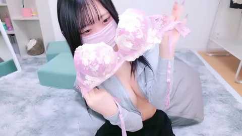 Media: Video of an Asian woman with long black hair, wearing a pink floral face mask and matching bra, sitting on a grey carpet in a modern living room.