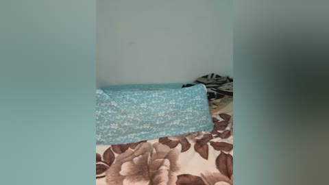 Media: A video of a child's bedroom corner featuring a turquoise pillow with white floral pattern on a beige bedspread with brown leaf designs. A white and black zebra-patterned pillow is slightly visible in the background. The room has a pale blue wall.