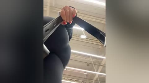 Media: Video of a person with light skin, wearing black leggings, pulling down the waistband, revealing a plump, curvaceous buttock. Background shows a warehouse ceiling with exposed pipes and fluorescent lights.