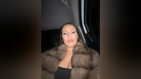 Media: Video of a woman with medium skin tone and straight dark hair, wearing a black top and a brown fur coat, sitting in a car, dimly lit.