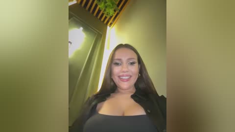 Media: Video of a smiling, dark-haired woman with long straight hair, wearing a black top that reveals her cleavage, in a modern, green-painted hallway with wooden accents and a green potted plant.