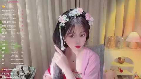 Media: Video of an East Asian woman with long black hair adorned with a floral crown, wearing a red dress with a low-cut neckline, seated indoors, surrounded by a cozy room with beige curtains, a lamp, and a small table.