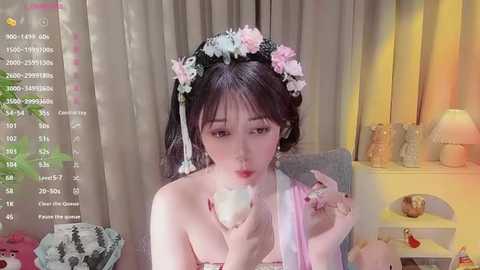 Media: Video of an Asian woman with pale skin, wearing a pink flower crown, blowing a pink balloon, in a cozy room with curtains and a lamp.