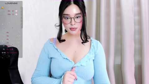 Media: Video of a young woman with long black hair, wearing glasses, a light blue off-shoulder top, and a microphone. She's seated indoors, with a calendar and curtains in the background.