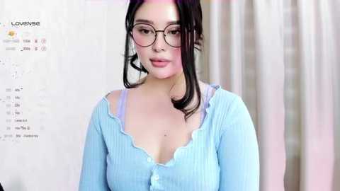 Media: Video of a young woman with fair skin and dark hair, wearing glasses and a light blue, ruffled blouse, against a blurred background with a \"Love\" sign.