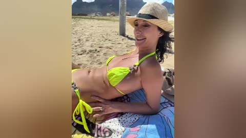 Media: Video of a curvy, light-skinned woman with curly hair, wearing a neon yellow bikini and straw hat, lying on a colorful beach towel on sandy beach.