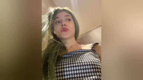 Media: Video of a young woman with long, wavy, green hair, wearing a black-and-white houndstooth dress. She has a light complexion and is indoors with neutral-toned walls. Her expression is neutral, and she appears to be in a casual, everyday setting.